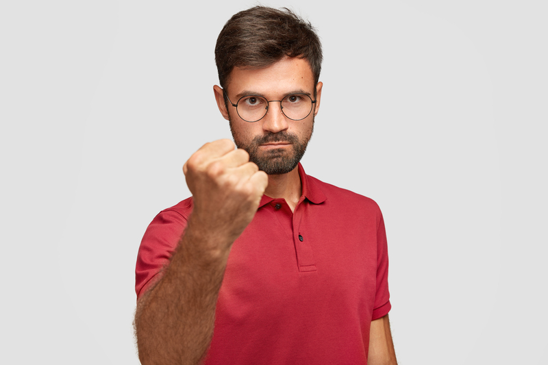 Serious angry male shows fist, ready for fight or challenge, has stern expression, wears casual red t shirt, poses against white background. Aggressive young man gestures indoor. Body language concept